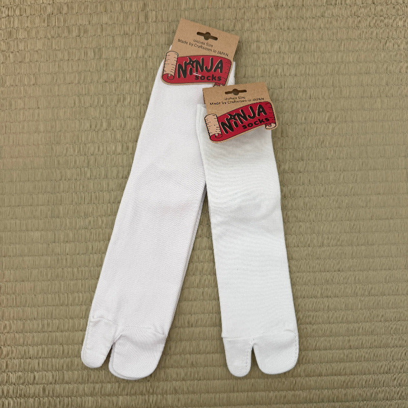A pair of white tabi-style split-toe socks with brown paper hanger tags reading 'NINJA SOCKS,' laid flat on a textured beige woven mat.