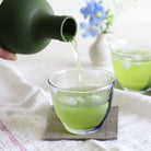 Green liquid being poured from a bottle into a glass on a white cloth background.