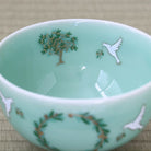 A light aqua ceramic bowl decorated with hand-painted green and gold olive tree motifs, white doves, and a green wreath with gold accents, shown on a woven beige mat.