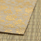 A close-up of a beige fabric or paper sheet with a metallic gold floral damask pattern, laid diagonally on top of a woven straw mat.