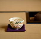 Crane and Sho-Chiku-Bai Kyo Ware matcha bowl with gold leaf accents displayed on a purple cloth in a Japanese tea room setting