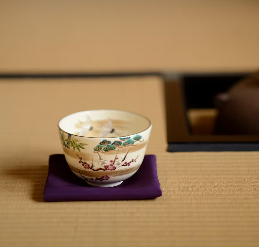 Crane and Sho-Chiku-Bai Kyo Ware matcha bowl with gold leaf accents displayed on a purple cloth in a Japanese tea room setting