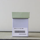 A small rectangular product box with a pale sage green lid and a white lower section, shown standing upright on a textured surface against a plain light background. A barcode label with Japanese text is printed on the front.