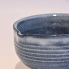 Angled side view of the denim-blue Tamba Ware matcha bowl showing its small pouring spout, ridged exterior, and softly varied glaze.