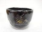 Front view of the Eiko Meido matcha bowl showing its softly irregular rim, smoky black glaze, pale markings, and gold botanical brushwork.