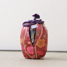 A small decorative ceramic vessel wrapped in a red floral fabric cover with a purple braided cord tied at the top, photographed against a plain light background.