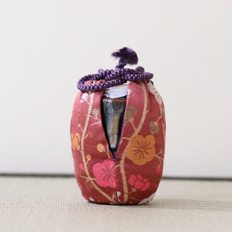 A small decorative ceramic vessel wrapped in a red floral fabric cover with a purple braided cord tied at the top, photographed against a plain light background.