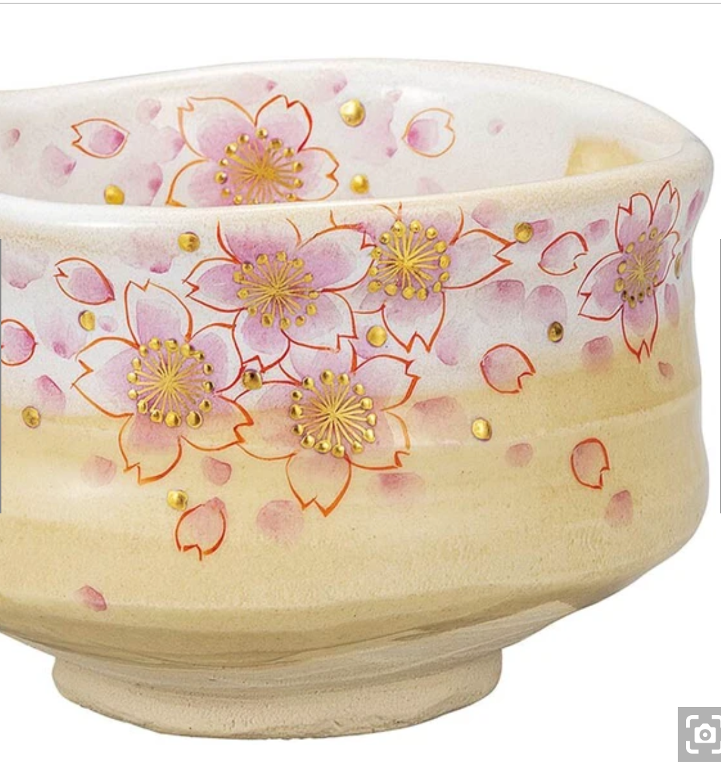 Left angled view of the Kutani Ware matcha bowl showing layered pink sakura blossoms, scattered petals, and gold details continuing around the exterior.