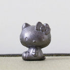 A small metallic Hello Kitty figurine with a textured silver finish, sitting on a light beige woven surface against a plain light gray background.