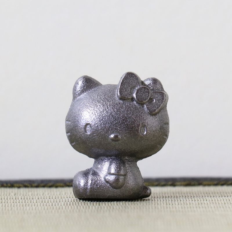 A small metallic Hello Kitty figurine with a textured silver finish, sitting on a light beige woven surface against a plain light gray background.