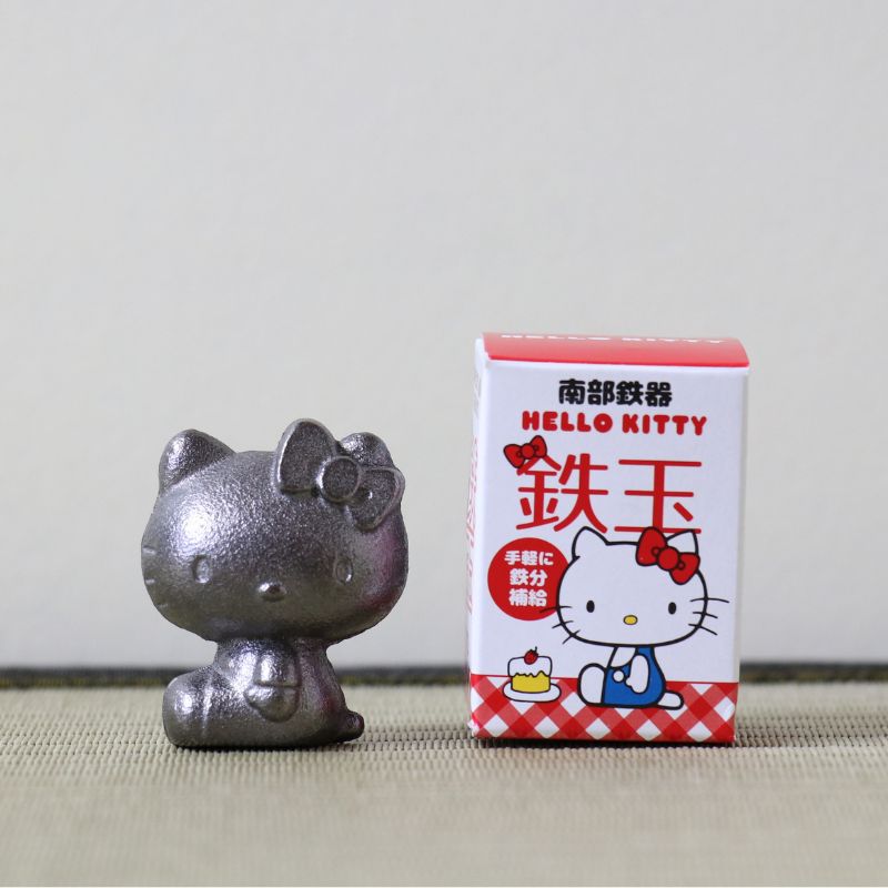 A metallic silver Hello Kitty figurine sits next to its original white and red retail box on a neutral beige surface against a light gray background.