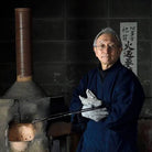 An older man wearing a dark blue traditional robe and white gloves stands beside a large wood-burning stove, holding a long metal tool. A black tiled wall and a vertical calligraphy sign are behind him.