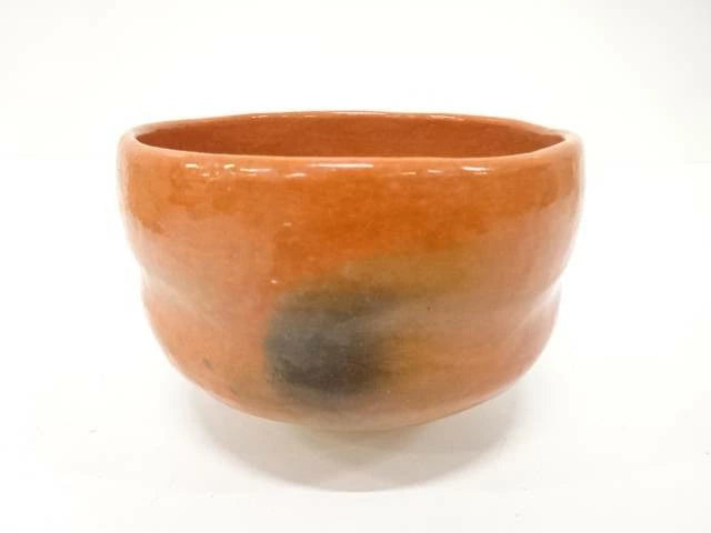 Front view of the Juraku Aka Raku chawan showing its warm coral-red glaze, rounded handcrafted form, and smoky black markings.