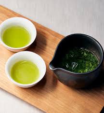 Two small white bowls with green liquid and a black teapot on a wooden board.