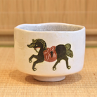 Front view of the white Raku matcha bowl showing the running black horse motif and softly irregular cylindrical form.