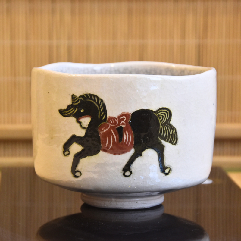 Main front view of a Kawasaki Waraku Shiro Raku matcha bowl with a hand-painted black horse, red decoration, and gold-toned details on a cream-white glaze.