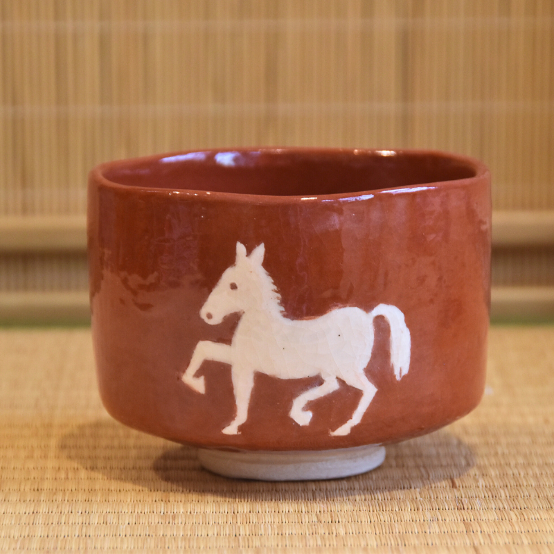 Front view of the red Aka Raku matcha bowl showing the white horse motif and softly irregular cylindrical form.