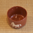 Angled top view showing the warm red interior of the Aka Raku matcha bowl and part of the white horse decoration.