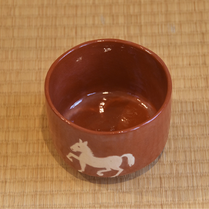 Angled top view showing the warm red interior of the Aka Raku matcha bowl and part of the white horse decoration.