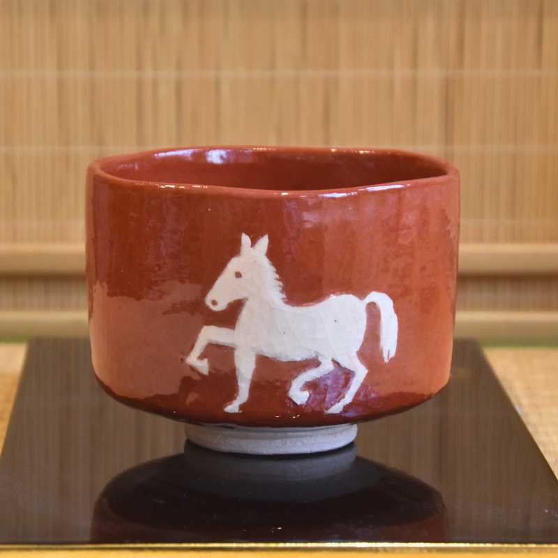 Main front view of a Kawasaki Waraku Aka Raku matcha bowl with a hand-painted white horse on a warm red glaze.