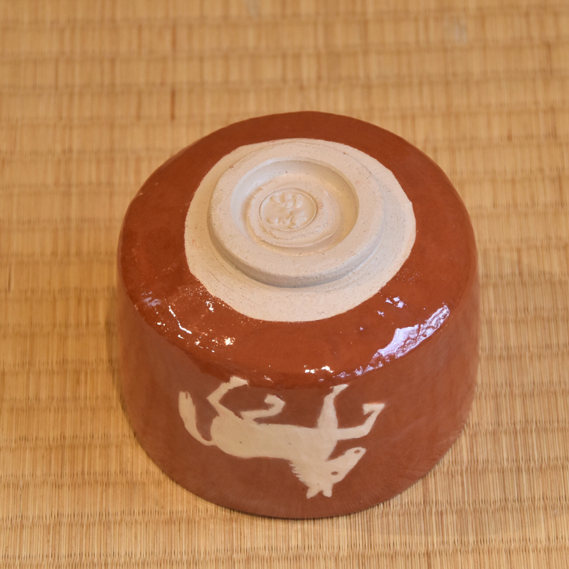 Underside angle of the red Raku matcha bowl showing its pale exposed foot and white horse motif wrapping around the exterior.