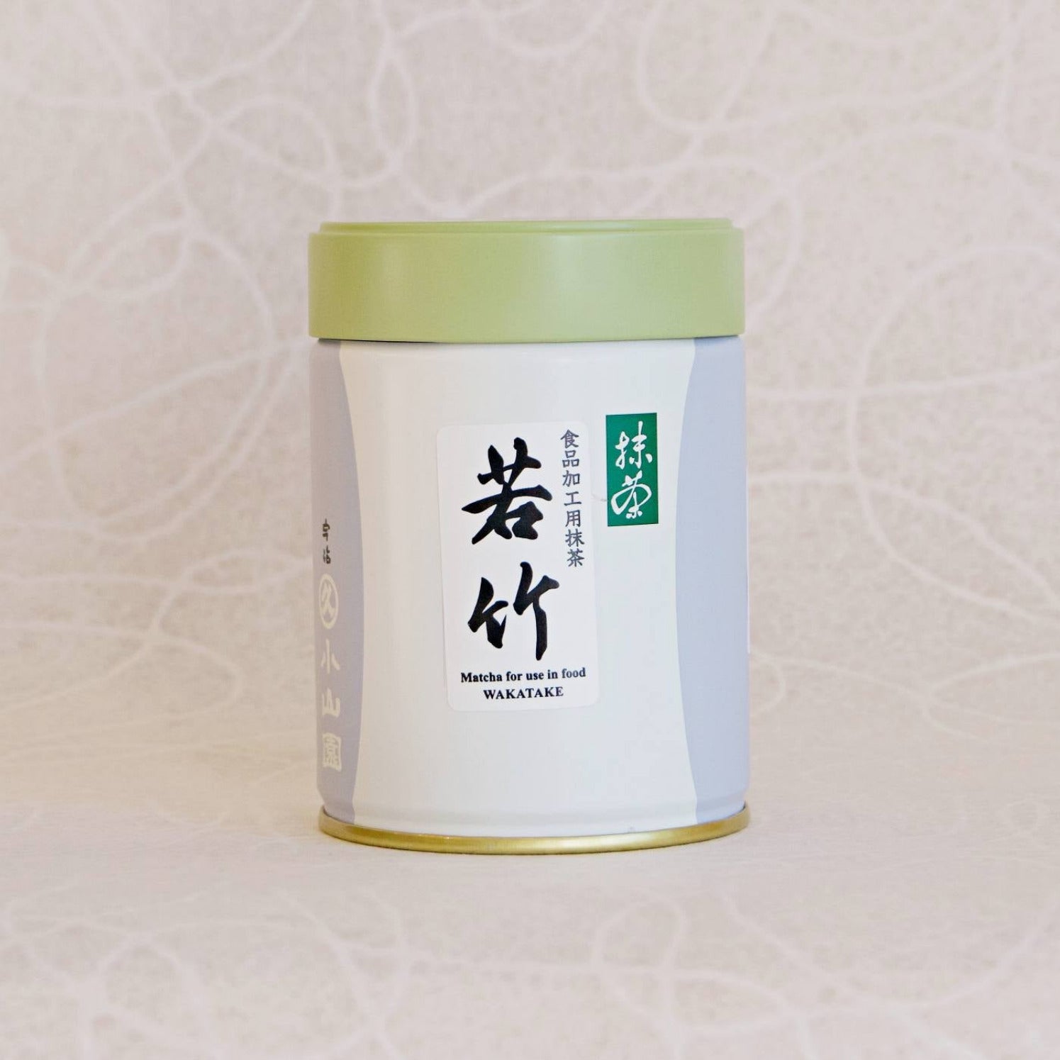 A can of TOMOTAKE Marukyu Koyamaen Matcha powder, with a predominantly white label and green lid, placed against a patterned background.