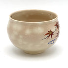 Side view of the hand-painted Kyo-yaki chawan showing red autumn maple leaves and curved blue flowing water against the softly mottled beige glaze