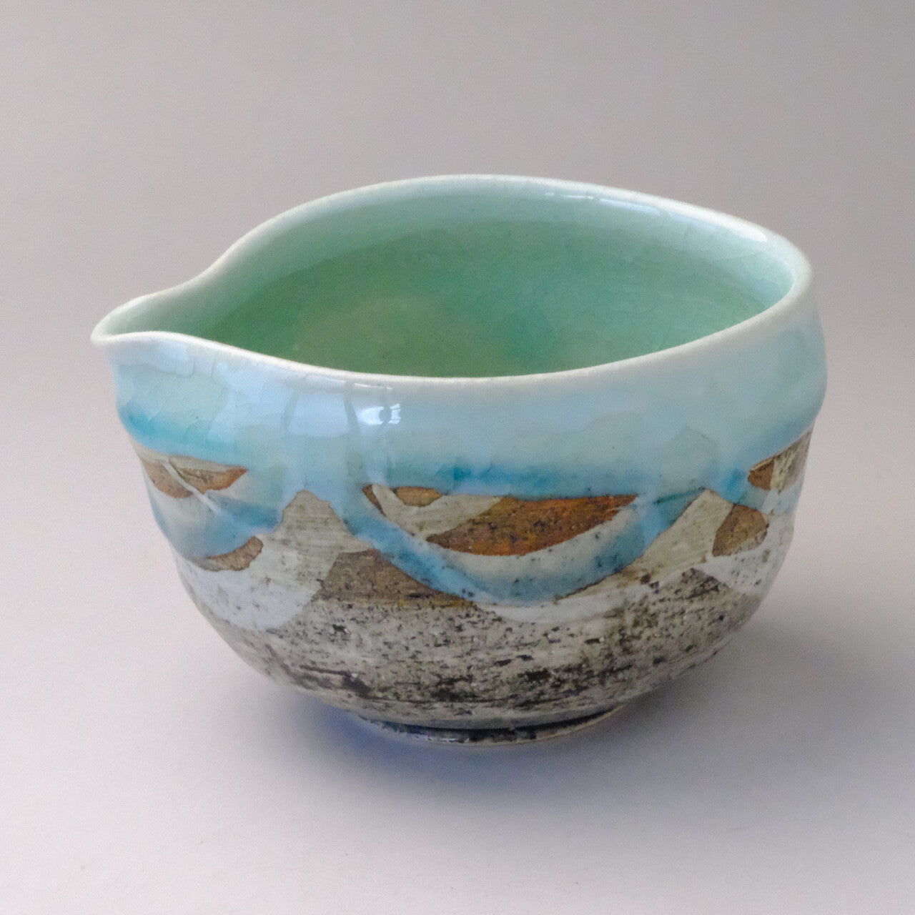Three-quarter view of a mint blue Mino Ware katakuchi matcha bowl with a pouring spout and flowing blue, white, and earth-tone glaze.