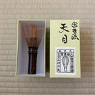 A traditional Japanese bamboo whisk (chashaku) with a dark brown handle, displayed next to its light green presentation box on a beige woven tatami mat background.