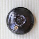 A round glossy black ceramic bowl with a spiral design on the bottom, placed on a light beige textured mat.