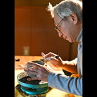 An elderly potter with gray hair and glasses works at a pottery wheel, shaping a small clay vessel with a tool. The warm, golden light illuminates the potter's focused expression and the clay, set against a softly blurred brown background.