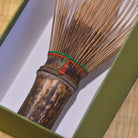 Traditional Japanese whisk with wooden black bamboo handle, cotton threads in red and green and thin bristles inside the green box with a light white inner background.