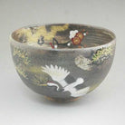 Front view of a hand-painted Sakata Yahata Kyo Ware matcha bowl with a large white crane, red and white plum blossoms, and gold-toned botanical accents on charcoal glaze.