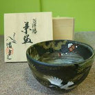 Sakata Yahata crane and plum blossom matcha bowl displayed with its box and accompanying papers against a green background.