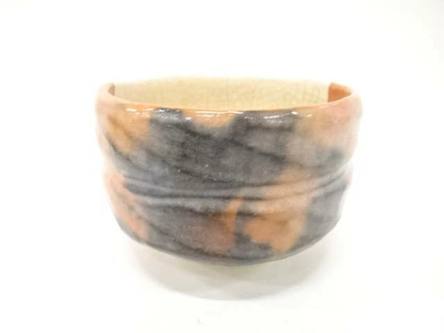 Side view of the Shoraku Sasaki Kotobuki Red Raku chawan showing its warm red-orange surface, hand-formed shape, and flowing dark charcoal glaze