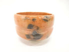 Front view of the Matsukaze Red Raku matcha bowl with persimmon-red glaze, smoky charcoal markings, horizontal ridges, and a pale foot.