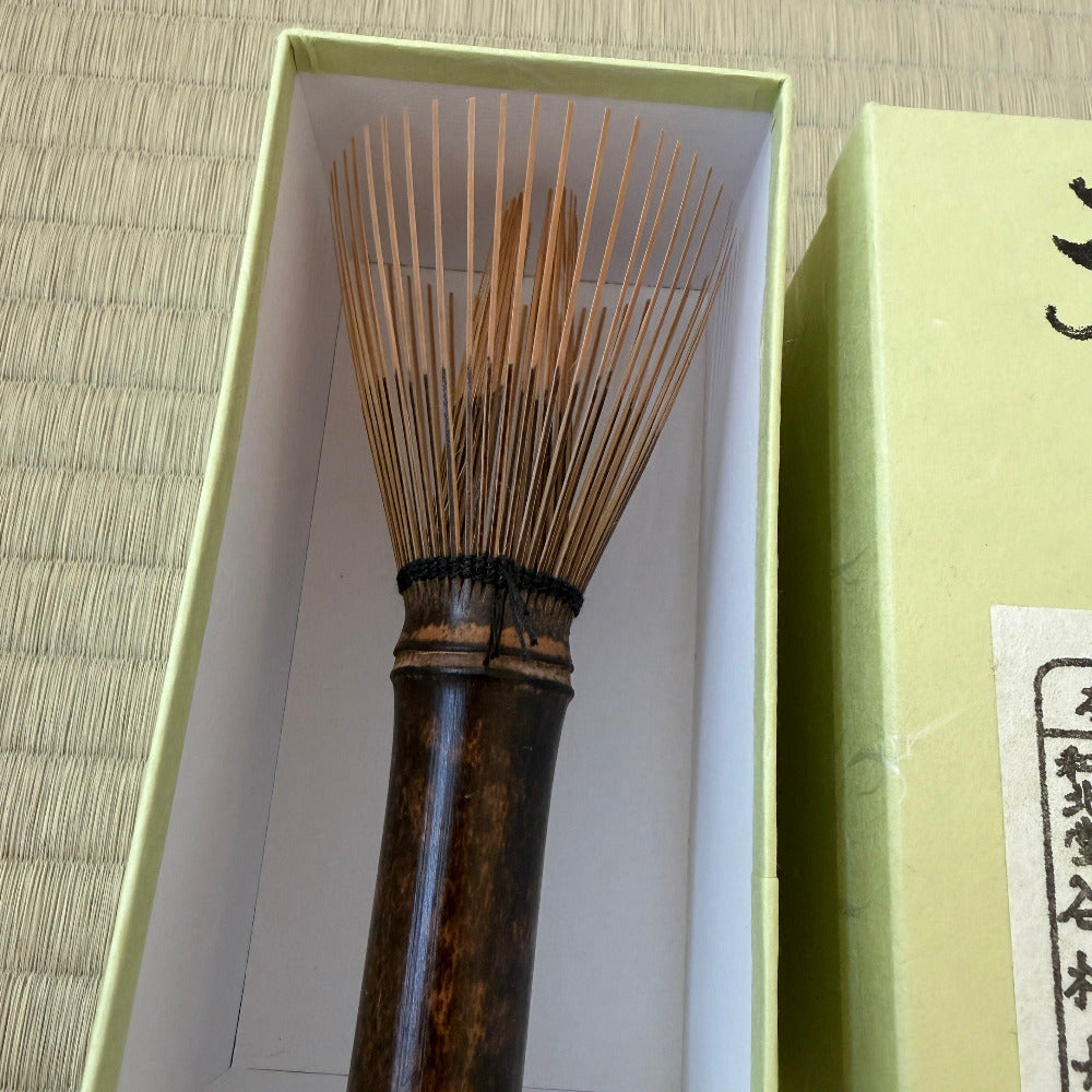 A traditional Japanese bamboo whisk (chashaku) with a dark brown handle and light tan bristles, presented inside its light green gift box next to its outer packaging on a tatami mat background.