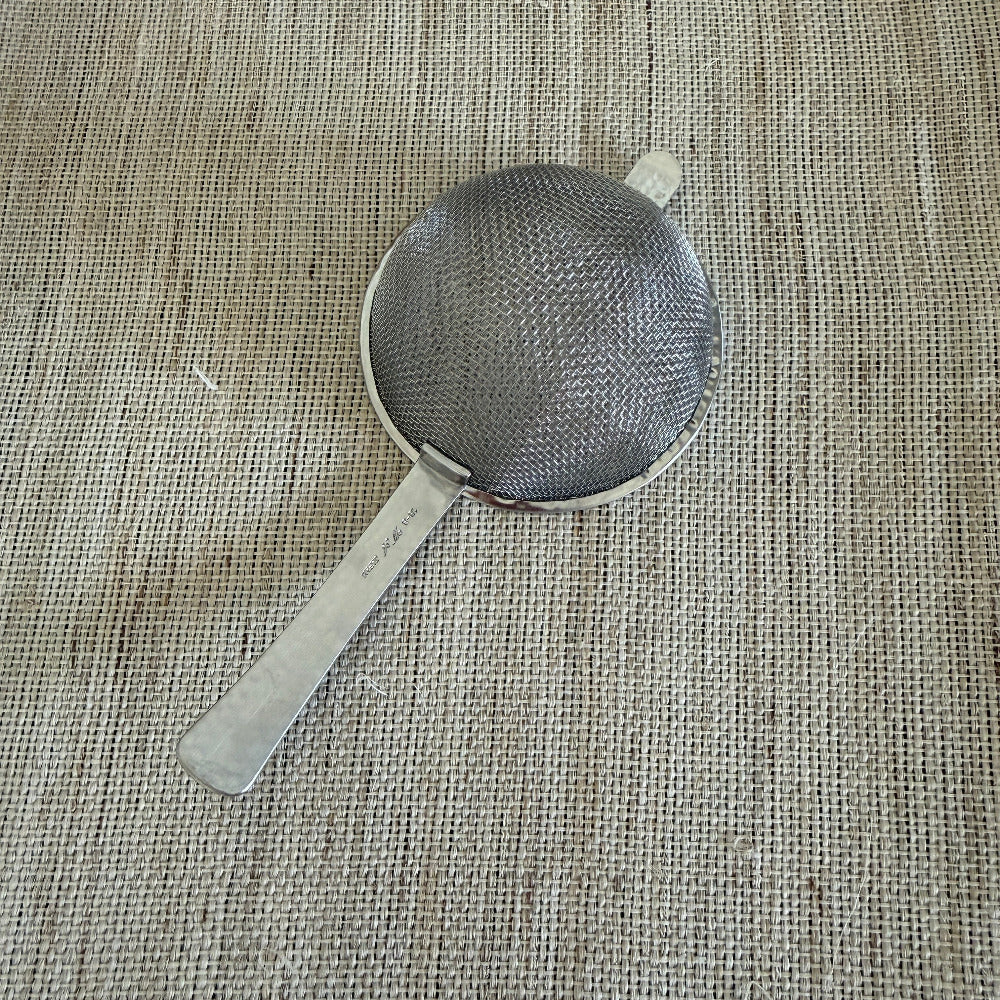 A small stainless steel mesh strainer with a flat handle, placed diagonally on a textured beige burlap fabric background.