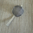 A small stainless steel mesh strainer with a flat handle, placed diagonally on a textured beige burlap fabric background.