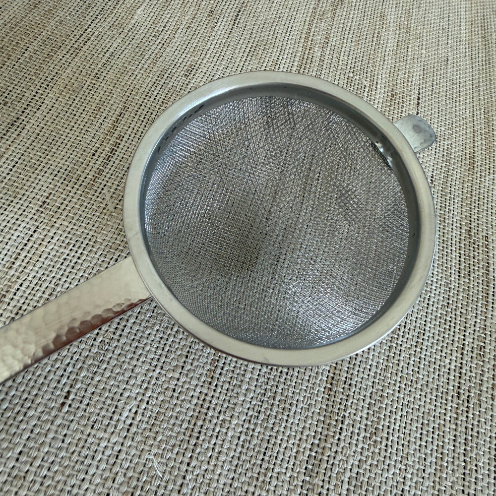 A round metal mesh strainer with a hammered finish on its handle, resting on a textured beige burlap surface.