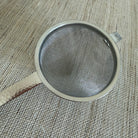A round metal mesh strainer with a hammered finish on its handle, resting on a textured beige burlap surface.