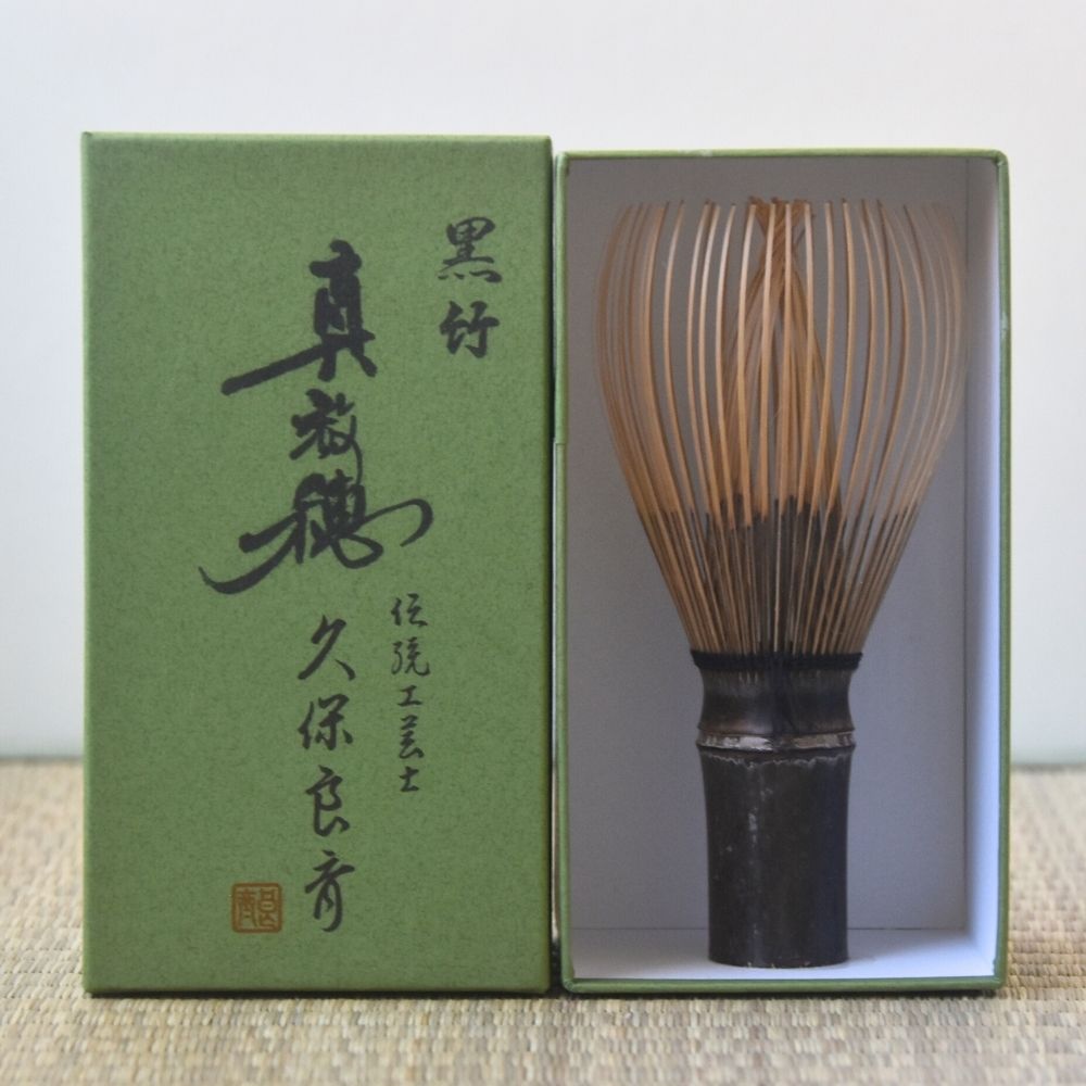 Traditional Japanese bamboo whisk with green box featuring Japanese text on a light background