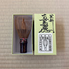A traditional Japanese bamboo matcha whisk displayed next to its light green presentation box on a beige woven straw mat background.