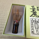 A bamboo and wood matcha whisk with a colorful cord tie, presented inside a light green gift box next to its lid on a woven mat background.