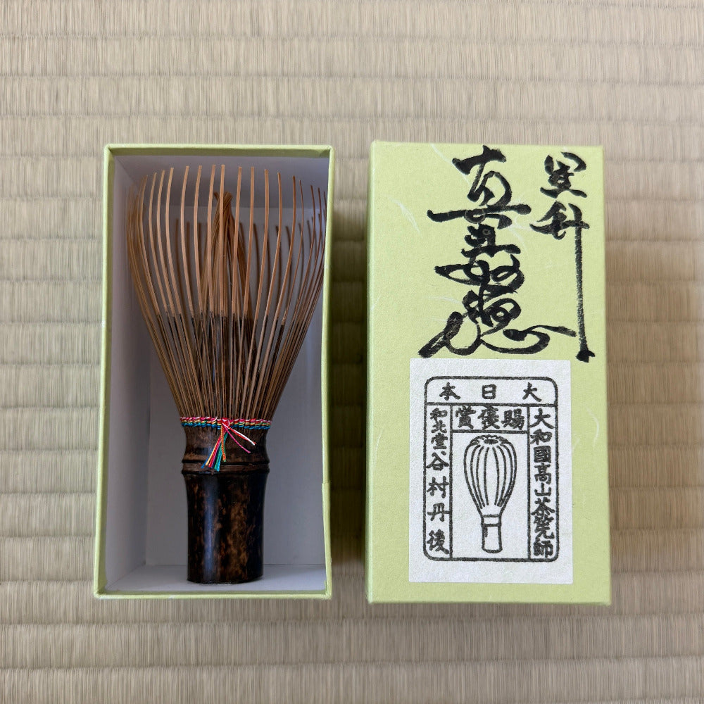 A traditional Japanese matcha whisk (chashaku) with a dark brown handle and bamboo tines, displayed next to its light green packaging box on a beige woven mat background.