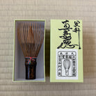 A traditional Japanese matcha whisk (chashaku) with a dark brown handle and bamboo tines, displayed next to its light green packaging box on a beige woven mat background.