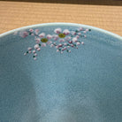 Close-up of the turquoise matcha bowl showing delicate pink and white cherry blossoms with green details near the rim.