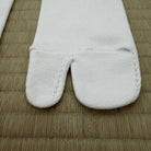 A close-up of a white tabi sock with its split toe separated, laid flat on a woven straw mat.