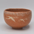 Front view of a Yoshimura Raku-nyu Aka Raku-style matcha bowl decorated with white flying cranes on a warm reddish-orange textured surface.